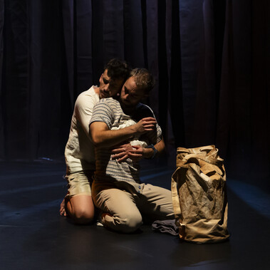 A tender scene. Two male presenting dancers are kneeling on the floor, one in front of the other, arms wrapped tightly around each other’s chest. It is Mariposa and the sailor Preston. Is this the start of a passionate love affair? A duffle bag is positioned in the foreground of the shot.