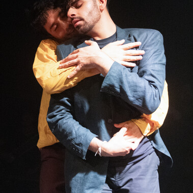Two male dancers, one in a light blue linen suit and the other in a yellow shirt with in a passionate embrace expressing love and happiness.