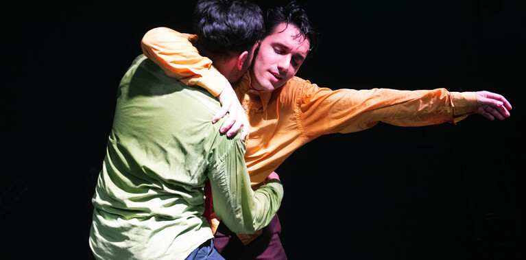 A male dancer in bright green shirt holding another male dancer in a bright yellow shirt in a tight embrace, with the latter’s arm outstretched expressing a passion of their desires..