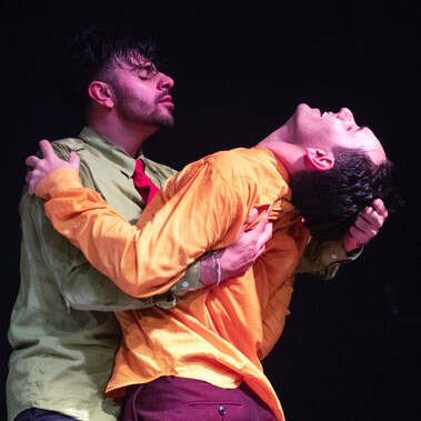 A male dancer in a light green shirt embracing another male dancer in a yellow short - pulling the latter’s head back by his hair. Their eyes are closed, expressing passion and desire of each other – confident in expressing their relationship.