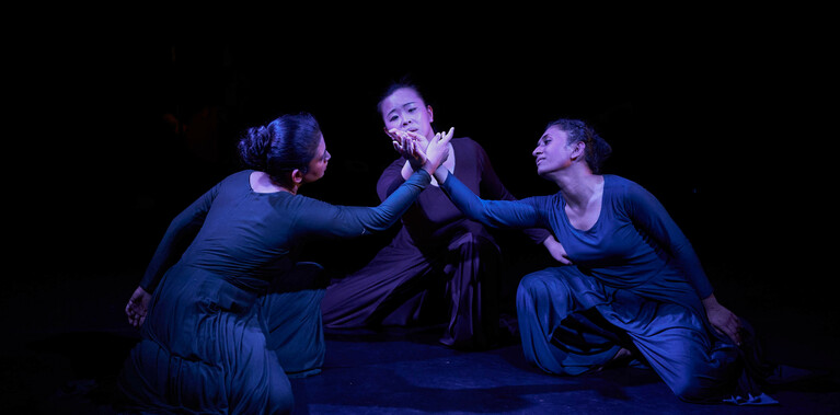 There are three dancers kneeling on the floor in a circle forming a single flower with their palms in solidarity and unity.