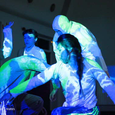 Lîla dancers dressed in white dance in a group. Blue and green light is projected on them.