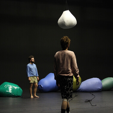 An image with a black background and black stage floor containing multiple large balloons of various colours (white, green, blue). You can see a male performer with his back facing us, carrying two balloons on either shoulder and only the arm and hand of another performer lying behind another balloon on the stage.