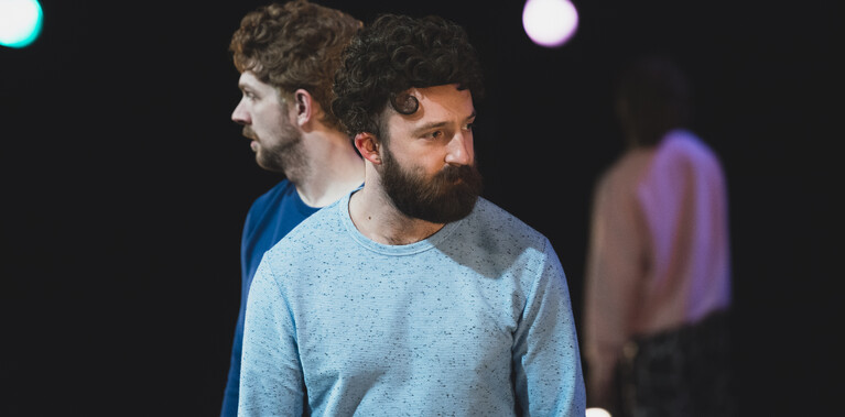 A close up shot of three male performers, two standing side by side yet facing looking outwards from one another and one facing away from us looking to the ceiling from a blurry distance.