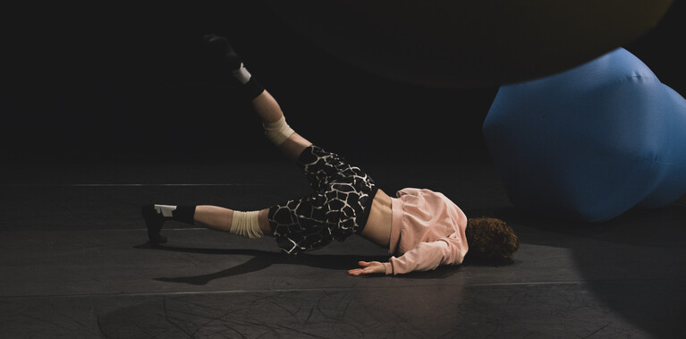 n image with a black background and black stage floor containing one blue balloon on the floor and one green balloon hanging from the ceiling. You can see a male performer in the centre on the floor, lying on his side with his back facing us. He has his left leg stretched in the air and is bearing weight on his shoulder.