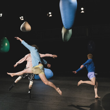 An image with a black background and black stage floor containing multiple large balloons of various colours (white, green, blue) with a string of stage lights surrounding the perimeter of the ceiling. You can see three male performers mid-air jumping with legs spread in a circle formation. You can not see any of their faces as they are either blocked by one another or facing away from us.